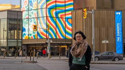 A person with short dark hair stands in front of her bright artwork on the front of the National Arts Centre building.